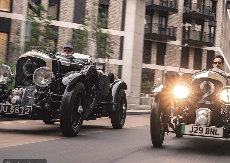 Two vintage racing cars driving side by side down a city street