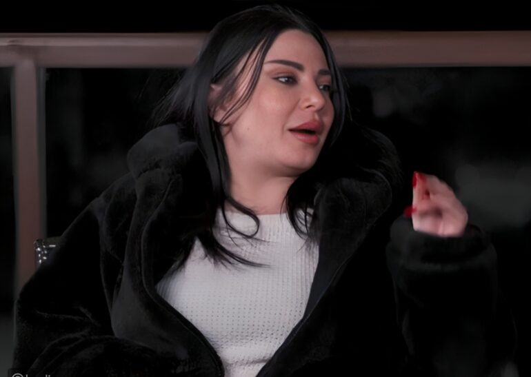 A woman in a dark coat reacting mid-scene inside a car